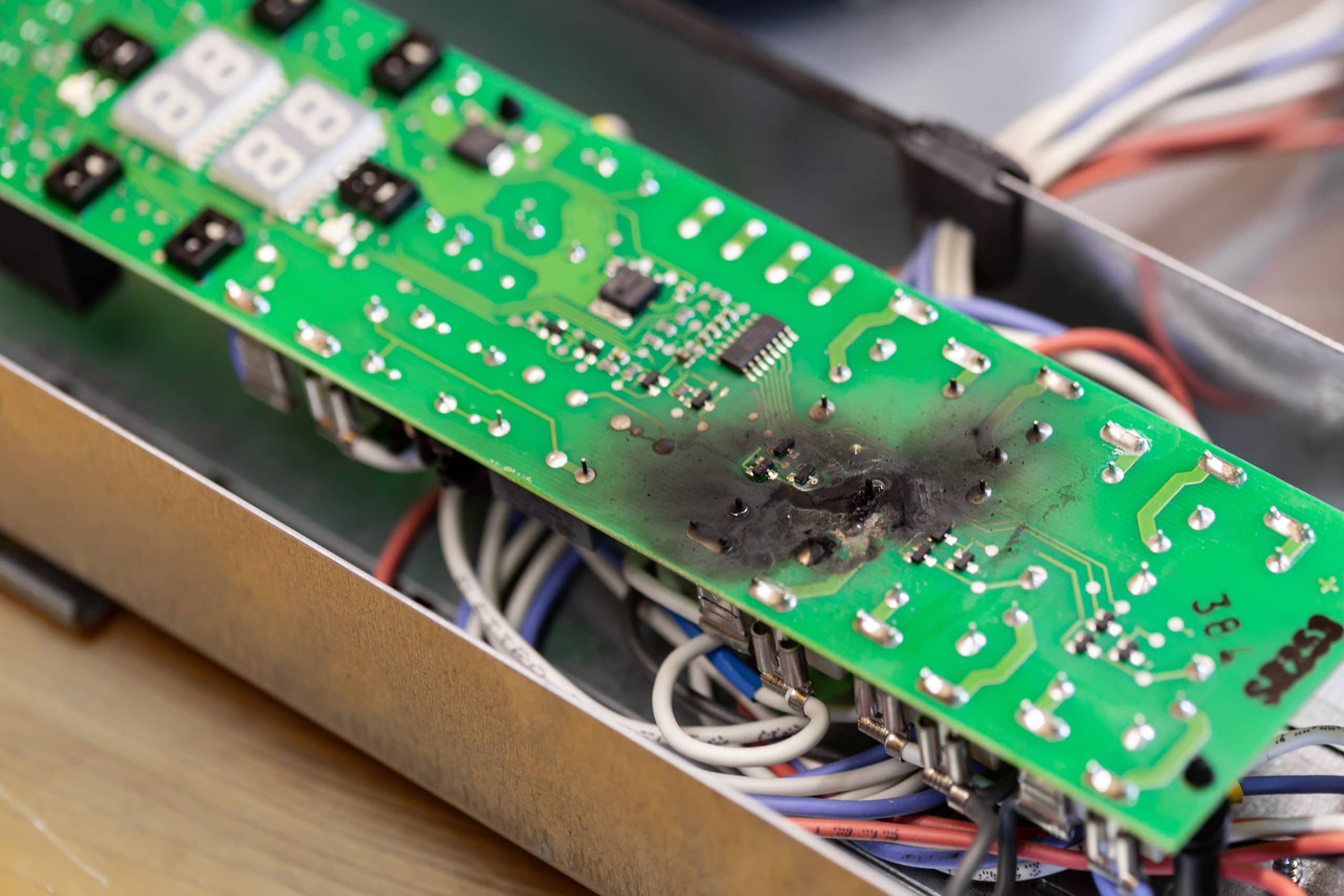 Burnt green PCB showing signs of electronic failure in metal casing with tangled wires.