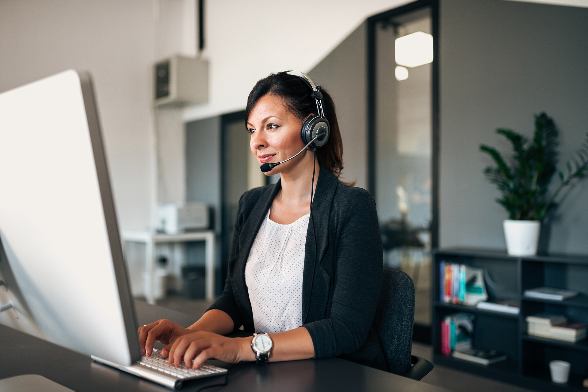 professional woman in modern office with headset, focused on computer screen in well-lit workspace.